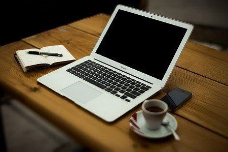 A laptop computer is sitting on a wooden table next to a cup of coffee and a notebook.